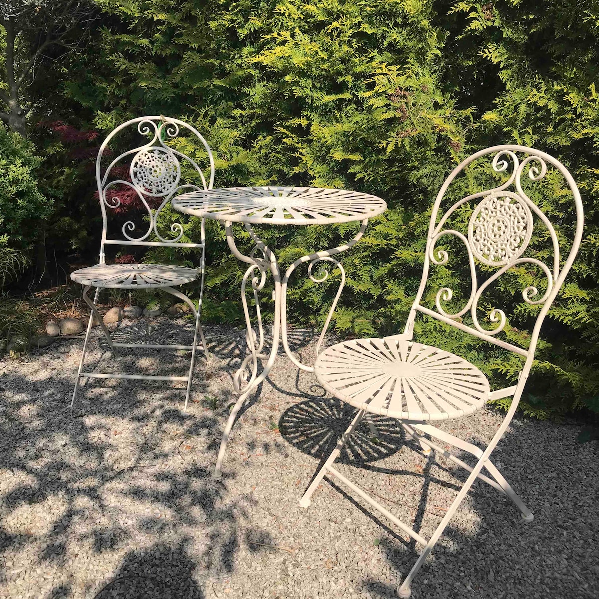 A quaint outdoor seating area is presented, featuring a small round table surrounded by two elegantly designed metal chairs. The setting is complemented by lush greenery in the background, creating a peaceful environment suitable for relaxation.