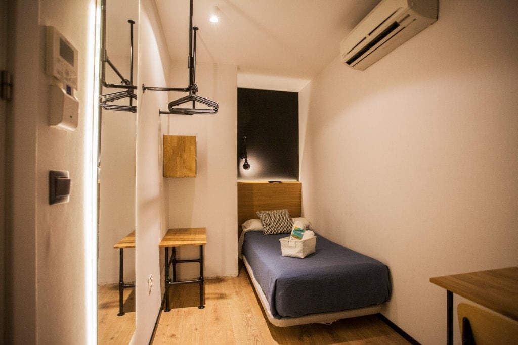A simple room is shown with a single bed dressed in a blue coverlet. A wooden nightstand and a desk are positioned beside the bed. Minimalistic decor is highlighted by warm wooden flooring and a wall-mounted air conditioning unit. Natural light enters through a window covered by a dark curtain.
