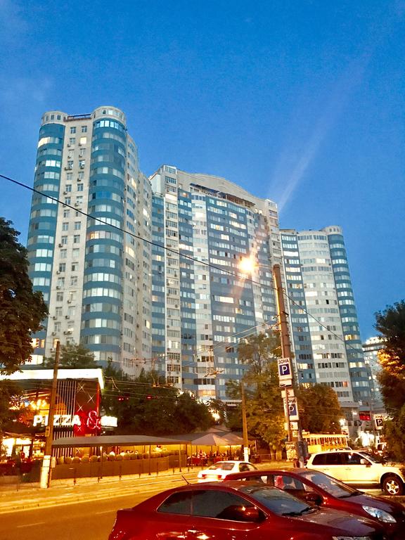 A modern high-rise building with a unique architectural design is showcased against an evening sky. The structure features a mix of glass and concrete, highlighting numerous windows that reflect the ambient city lights. Surrounding trees and street lamps create a lively urban atmosphere.