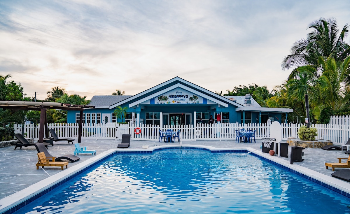 A large swimming pool sits at the forefront, reflecting the clear sky. A spacious building with a blue exterior features numerous windows and doors, surrounded by tropical greenery. Lounge chairs are positioned on the pool deck, providing a relaxing area for guests.