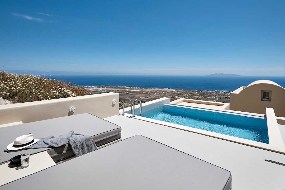 A sunlit terrace features two spacious loungers beside an outdoor heated jacuzzi. The view extends over the sea, showcasing a clear blue horizon and distant land. The design incorporates natural stone elements and minimalistic decor, creating a serene and inviting atmosphere.