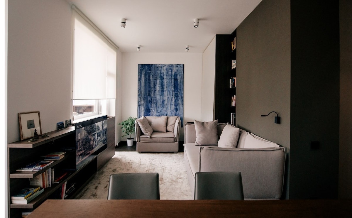 A modern living area features two comfortable sofas arranged around a neutral rug, with a large art piece in blue hues on the wall. Natural light pours in from large windows, and a bookcase is filled with various books. A small indoor plant adds a touch of greenery.