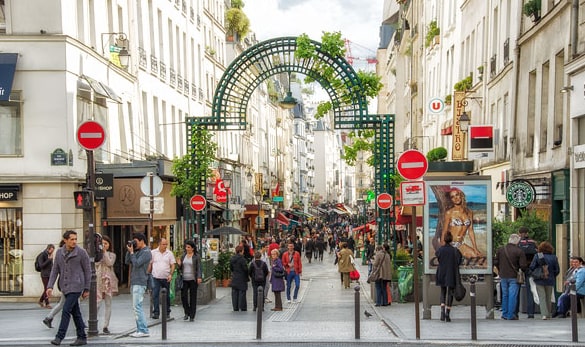 A lively pedestrian street in central Paris is displayed, flanked by various shops, cafes, and vibrant signage. An arch adorned with greenery marks the entrance, while crowds of people enjoy the bustling atmosphere. Sidewalks are lined with outdoor seating, inviting passersby to relax.