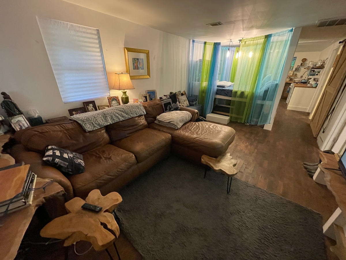A spacious living area features a large brown sectional sofa positioned on a soft gray rug. Natural light filters through partly sheer curtains, creating a warm ambiance. Decorative wood side tables and personal photographs enhance the inviting atmosphere, while a glimpse of the private sleeping area is visible in the background.