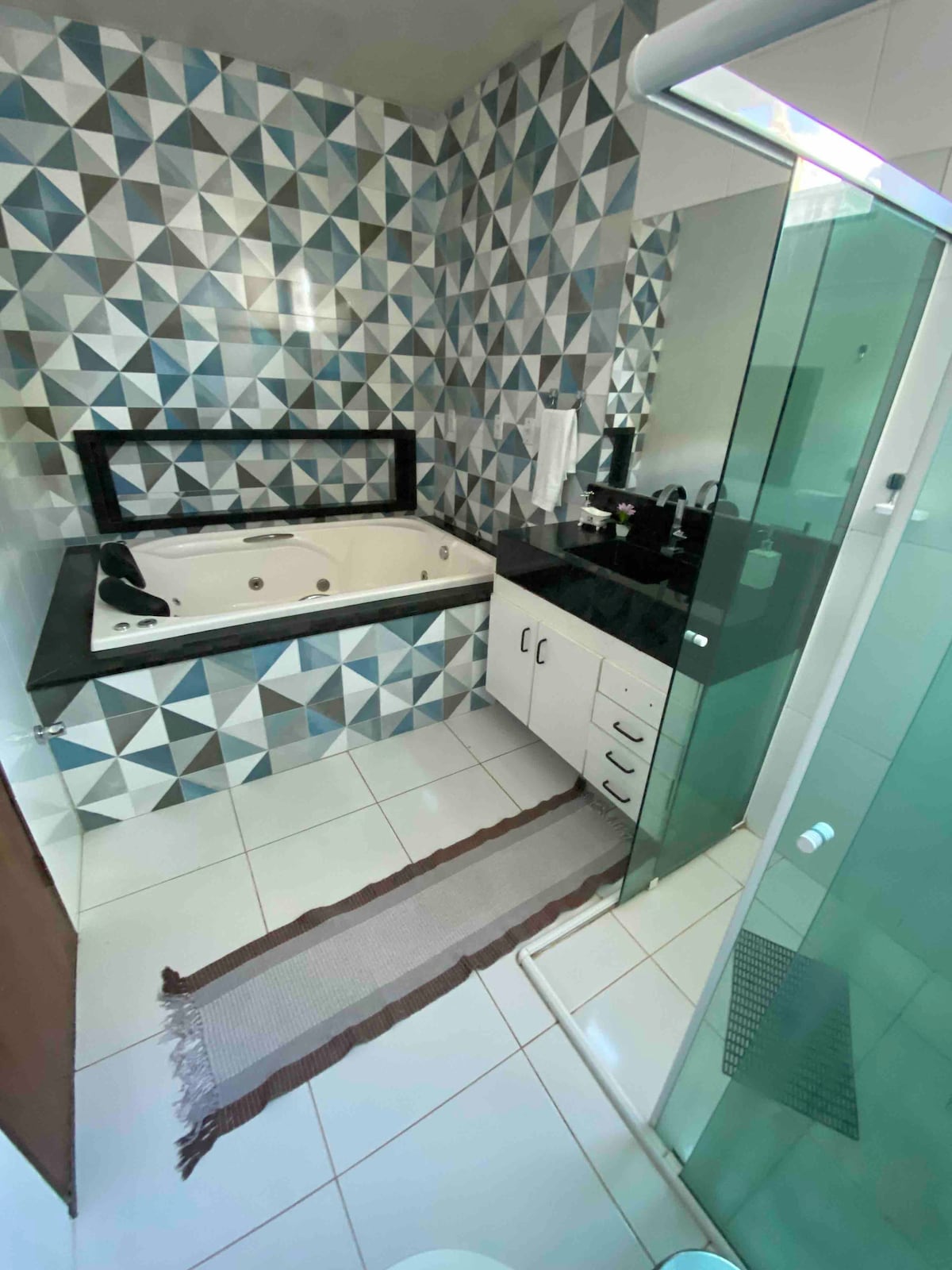 A spacious bathroom features geometric patterned tiles in shades of blue and white. A large whirlpool bathtub is positioned beside a glass-enclosed shower. Dark countertops are complemented by light cabinetry, and a soft rug lies on the tiled floor for added comfort.