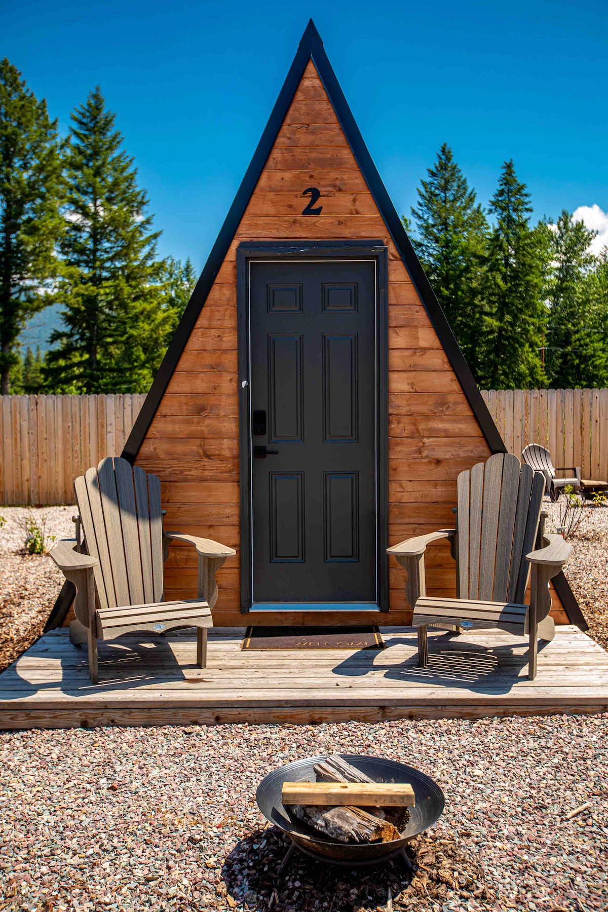 A cabin with a triangular roof is featured, showcasing a dark green door at the front. Two wooden Adirondack chairs are positioned on a small deck, with a circular fire pit containing firewood nearby. Surrounding trees and a privacy fence can be seen in the background.