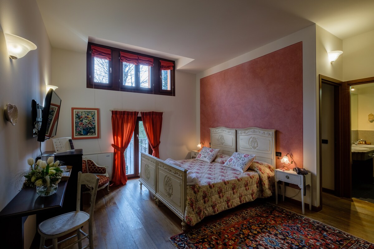 A spacious room features a floral-patterned bed with white wooden headboards. Natural light filters through three windows with red curtains. A wooden table with chairs is positioned nearby, and an adjoining bathroom is visible through an open door.