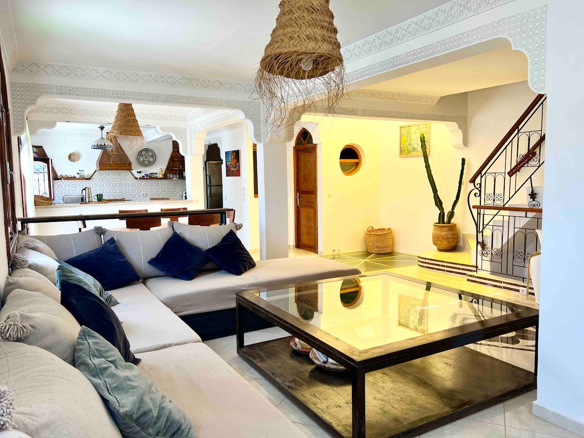A spacious living area is featured, characterized by a large, low coffee table surrounded by a comfortable seating arrangement. Natural light fills the space from a nearby window, highlighting textured decor and woven pendant lights hanging from the ceiling.