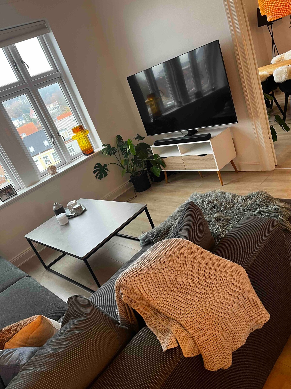 A cozy living room area features a comfortable gray sofa adorned with a soft, chunky-knit blanket. A minimalist coffee table stands in front of the sofa, accompanied by decorative items. A large flat-screen TV is mounted on the wall, while natural light enters from the window.