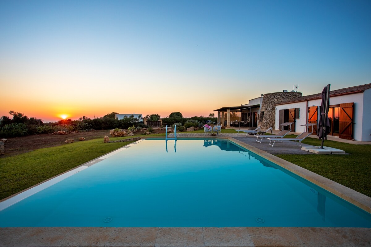 A serene sunset reflects off the clear waters of a private pool, framed by a well-kept lawn and gardens. The villa, with large glass doors, offers an inviting atmosphere. Comfortable lounge chairs are positioned nearby, enhancing the tranquil outdoor setting.