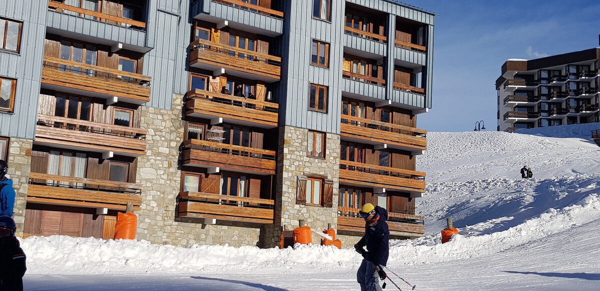 A multi-story building stands at the base of snow-covered slopes, featuring wooden balconies that overlook the area. Skiers navigate the terrain in the foreground, while orange barriers indicate designated areas. The backdrop includes additional buildings, enhancing the winter landscape.