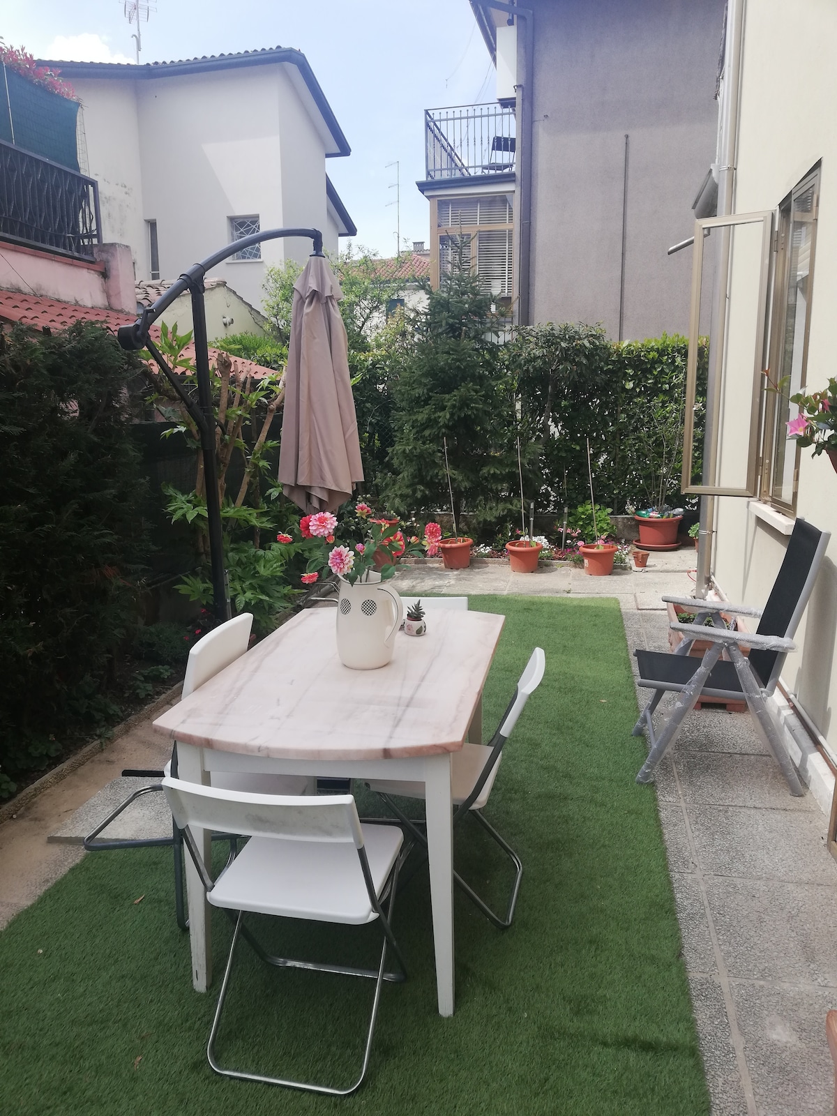 A garden area is presented with a rectangular table surrounded by four chairs on a green lawn. Flower pots are arranged along the sides, adding a touch of greenery. An umbrella is positioned above the table, providing shade. The space is bordered by neatly trimmed hedges and residential buildings.