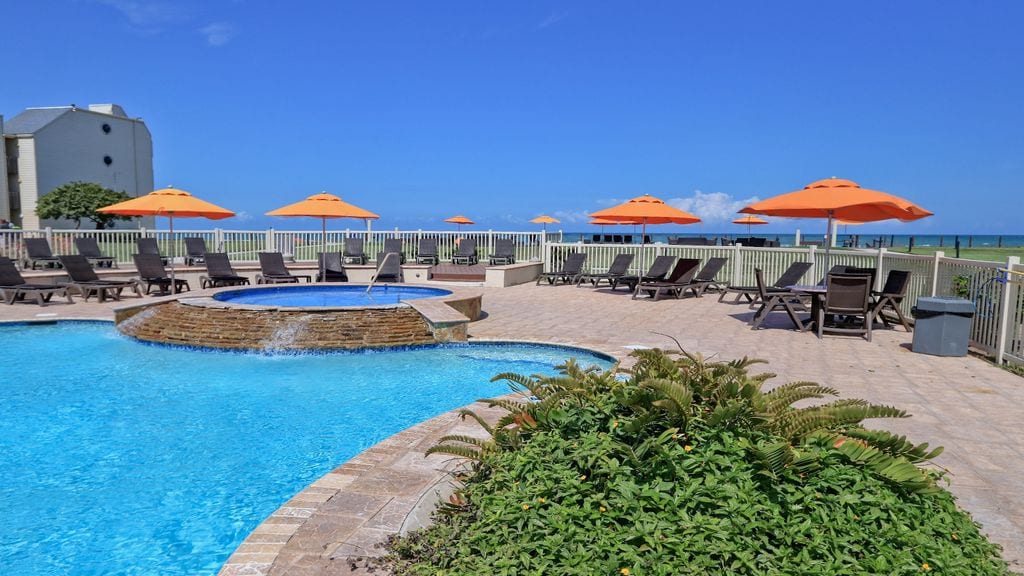 A tranquil pool area features a circular hot tub surrounded by vibrant lounge chairs. Bright orange umbrellas provide shade over the seating arrangement, while the sparkling blue water of the pool contrasts with the lush greenery nearby and the ocean view in the background.