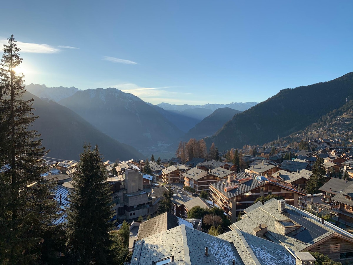 A view of the surrounding alpine landscape captures the valley and mountain ridges in the distance. The foreground features a mix of chalet-style buildings with sloped roofs, while sunlight filters through the trees, emphasizing the charm of the village.