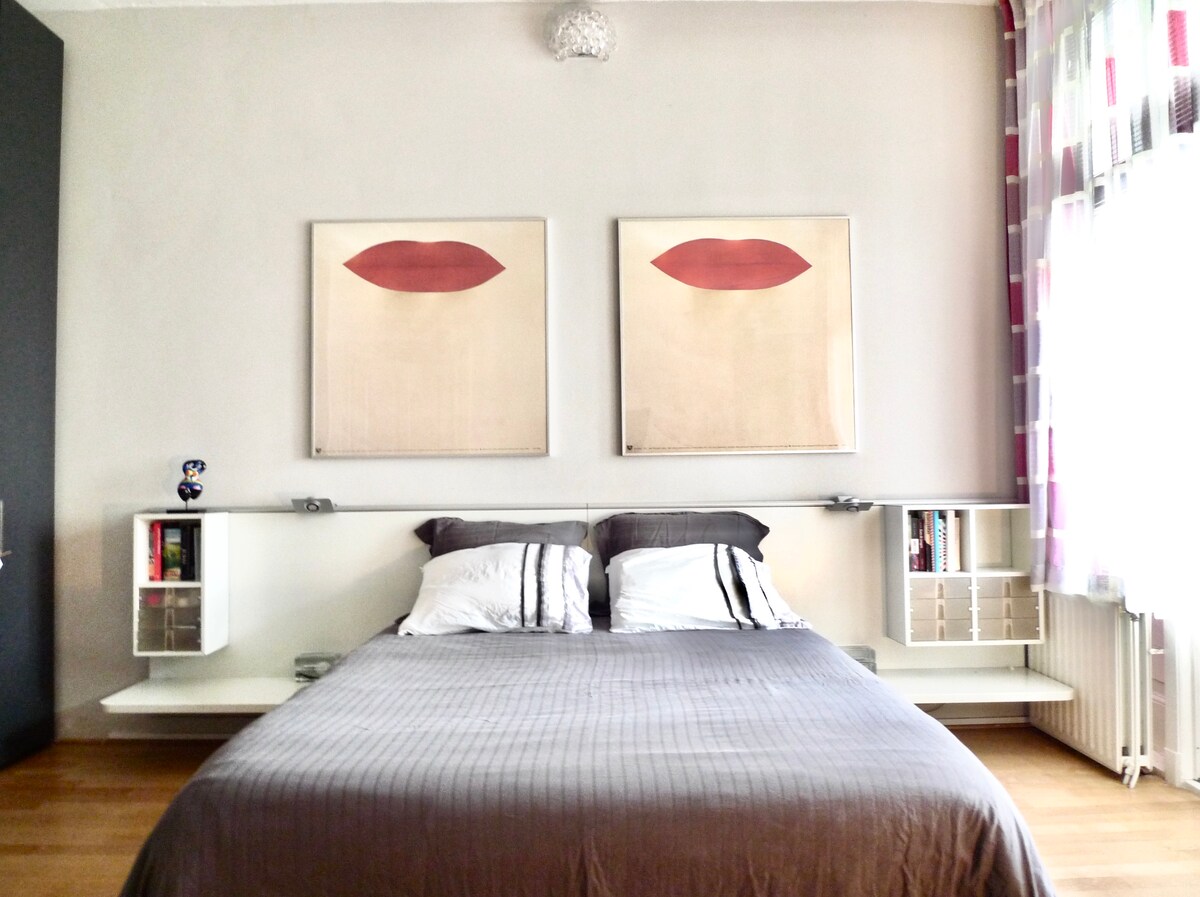 The master bedroom features a spacious bed with neatly arranged pillows and a grey duvet. Two framed artworks are displayed above the bed, depicting stylized lips. Modern bedside tables provide storage, complemented by natural light from the window adorned with patterned curtains.