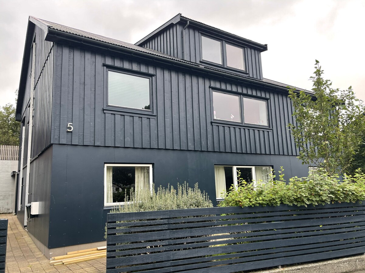 A modern, two-story building is presented with a deep blue exterior, featuring large windows that promote natural light. A neatly arranged fence enclosed with greenery frames the entrance, which is complemented by a simple pathway leading to the front door.