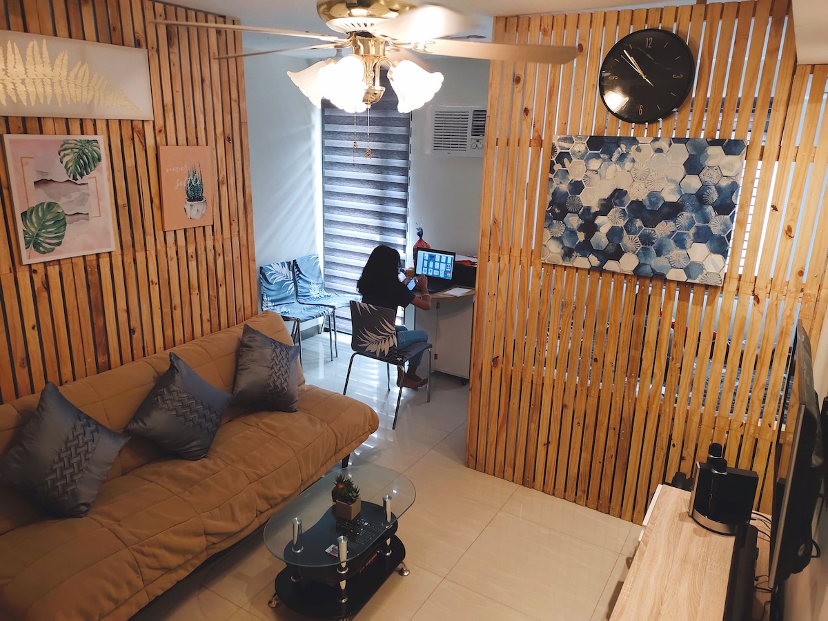 A compact living area features a beige sofa bed adorned with decorative cushions. A wooden accent wall complements framed wall art. A desk with a computer is visible, while a clock and large window with layered blinds add functionality and style to the space.