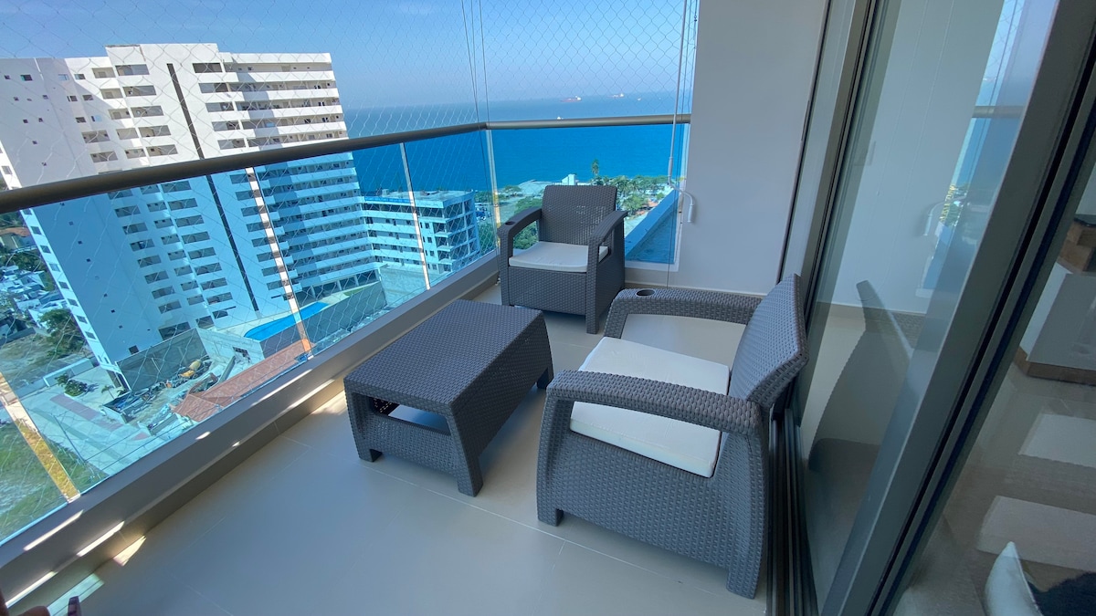 A balcony furnished with two armchairs and a coffee table offers views of the ocean and surrounding structures. The glass railing provides a clear sightline to the horizon, enhancing the sense of openness and connection with the landscape.
