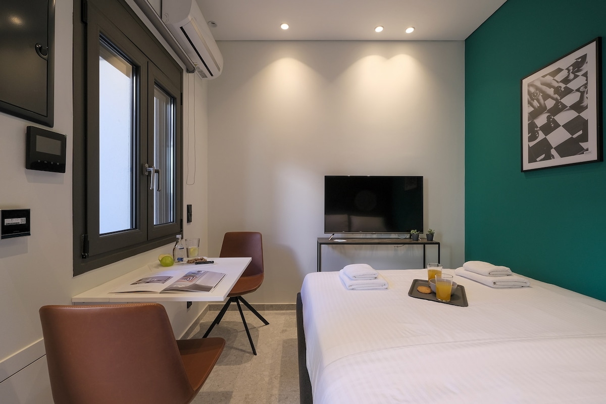 A bright bedroom features a spacious bed with fresh linen and towels placed neatly on top. A smart TV is positioned on a low table. A small dining area includes two chairs and a table, positioned near a window that allows natural light to fill the space.
