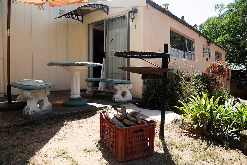 A shaded outdoor area features a stone table surrounded by two benches, providing a comfortable space for dining. A braai place with a heated grill is positioned nearby, along with a crate of firewood. Lush greenery and decorative plants enhance the natural surroundings.