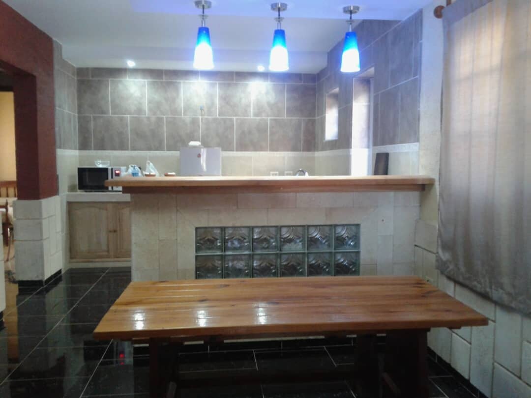 A modern kitchen features a wooden countertop with a glass-block bar area. Blue pendant lights illuminate the space, highlighting the sleek tile walls. A wooden table sits in the foreground, and windows allow natural light to enter, softened by light curtains.