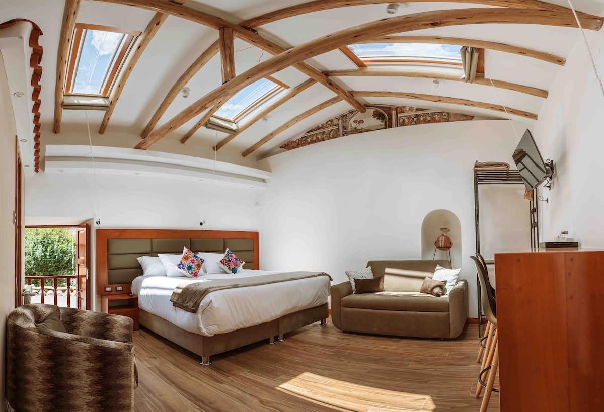 A spacious bedroom features a large bed adorned with colorful throw pillows. Natural light floods the room through skylights and a window, highlighting the wooden beams overhead. A cozy seating area includes a sofa, with a wooden desk and chair positioned nearby.