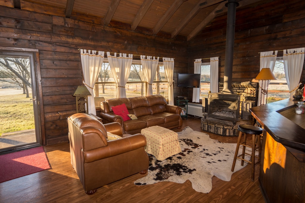 A spacious living area is highlighted by large windows that offer views of the outdoor scenery. A leather sofa and armchair are arranged around a central ottoman and a cozy fireplace. A television is positioned nearby, creating a comfortable and inviting space.