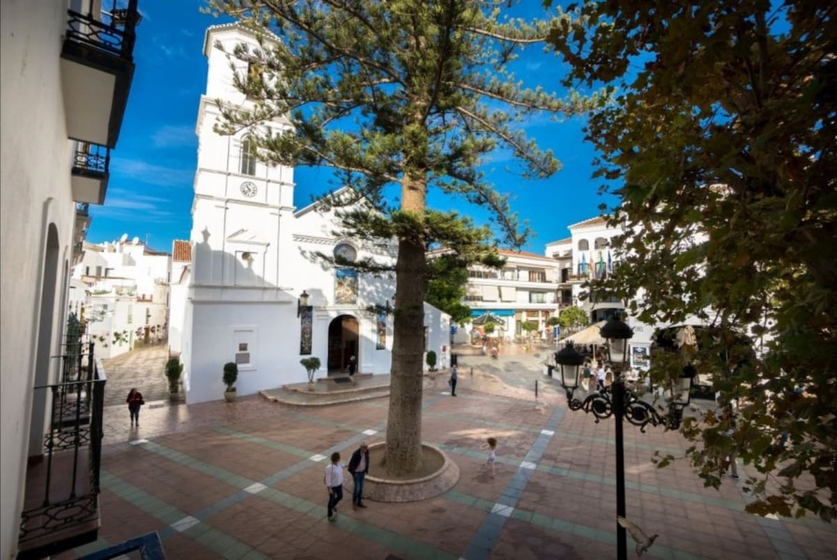 A vibrant town square is framed by charming white buildings, showcasing a notable church with a clock tower. A tall tree provides shade in the center, while pedestrians stroll along the paved walkway, indicating a lively atmosphere.