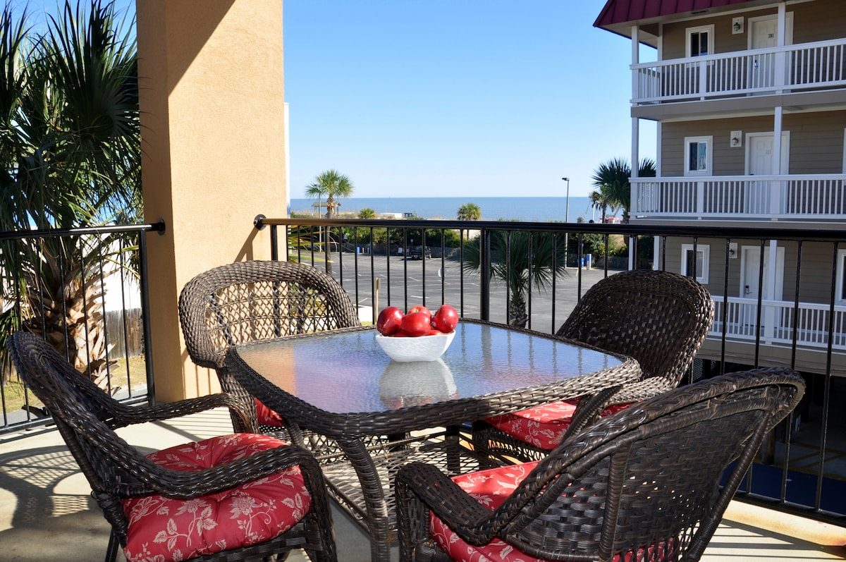 A covered balcony features a table surrounded by four wicker chairs with red cushions. A bowl of red apples sits at the center of the glass table. The view includes palm trees and the ocean in the distance under a clear blue sky.