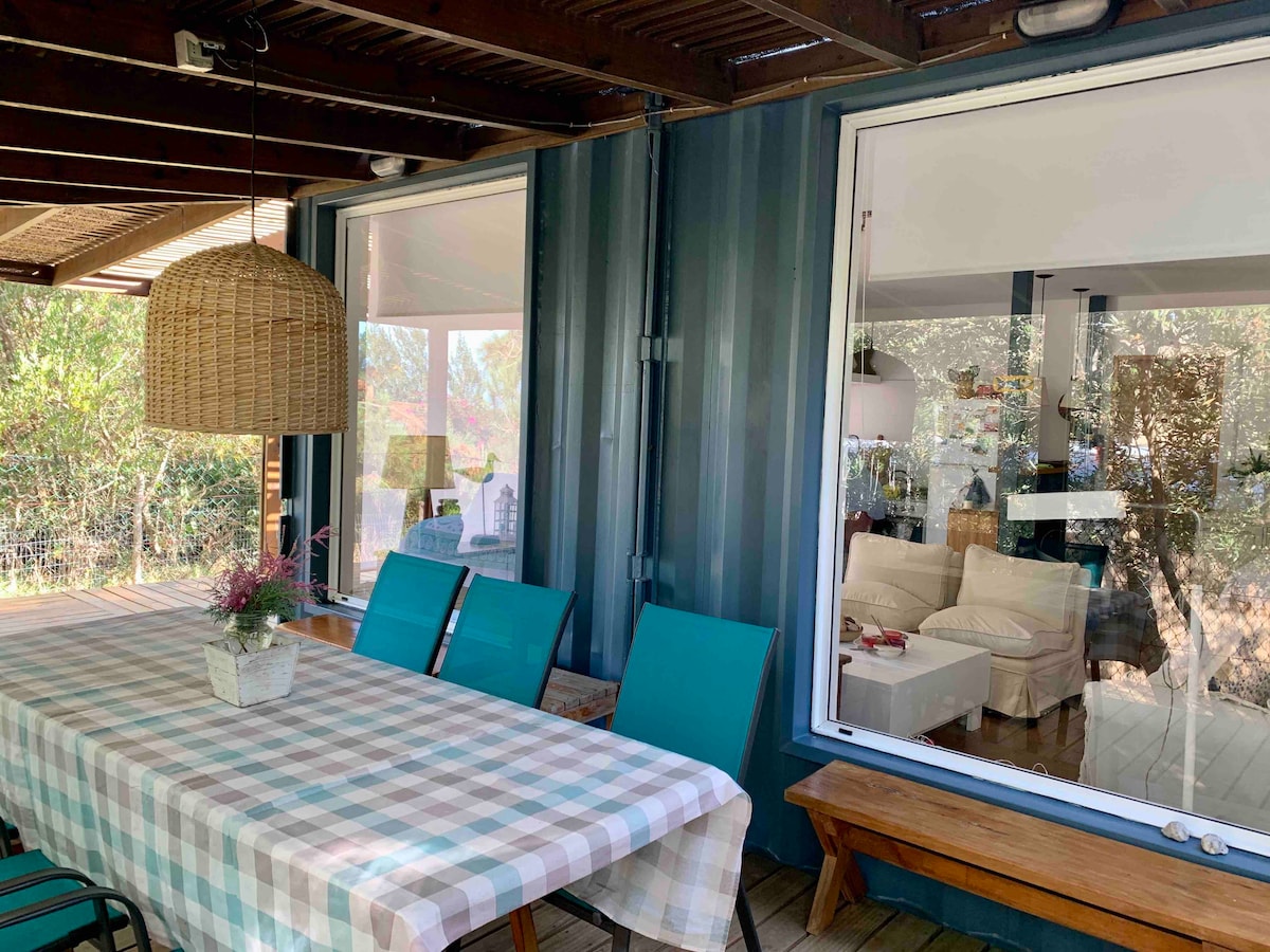 A covered outdoor dining area features a large table with a checkered tablecloth and several teal chairs. A hanging light fixture made of woven material is present above the table. Large windows provide a view into a bright living space with light-colored furniture.