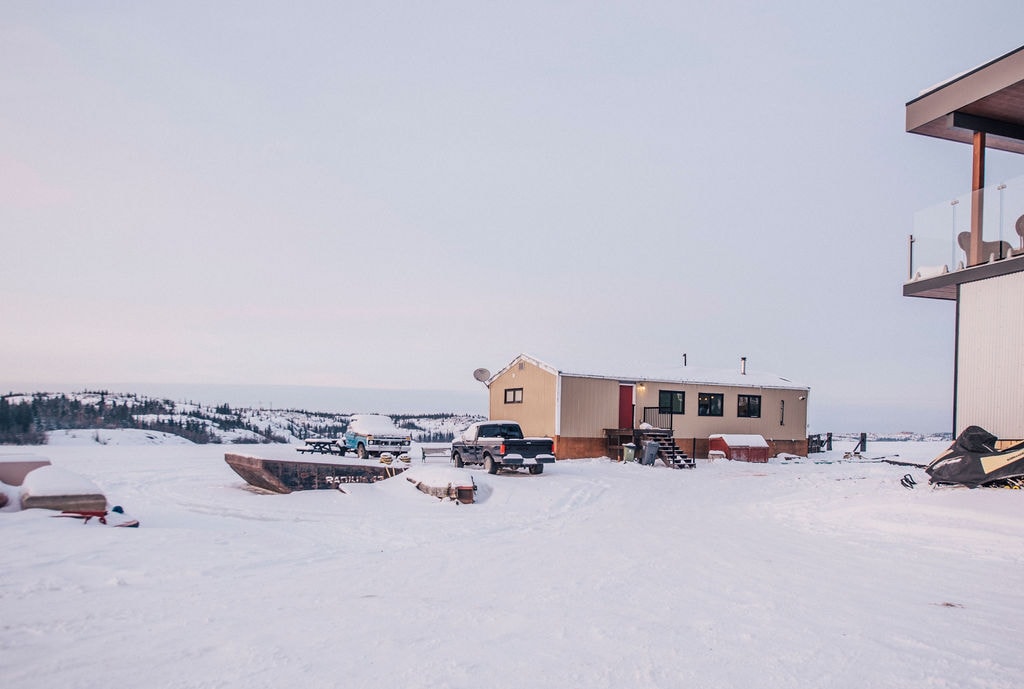 A winter scene showcases a serene waterfront property, with snow blanketing the ground. A structured building with a sloped roof is visible alongside multiple vehicles. The distant landscape features gently rolling hills and trees, under a soft sky with hints of color.