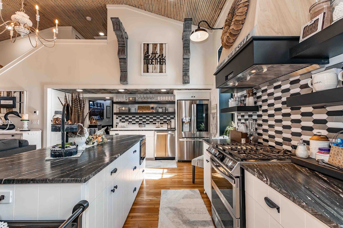 A gourmet kitchen features a central island with contrasting black and white cabinetry. Modern appliances, including a stainless steel refrigerator and professional-grade gas range, are present. The patterned tile backsplash adds visual interest, while natural light pours in from large windows, illuminating the space.