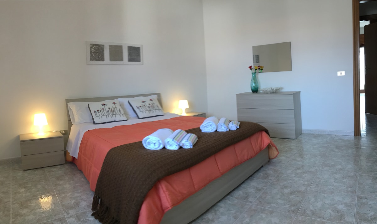 A well-lit bedroom features a spacious bed covered with an orange and brown throw. Two nightstands with lamps stand on either side of the bed. A dresser with a mirror is positioned against the wall, and decorative items are displayed on the dresser.