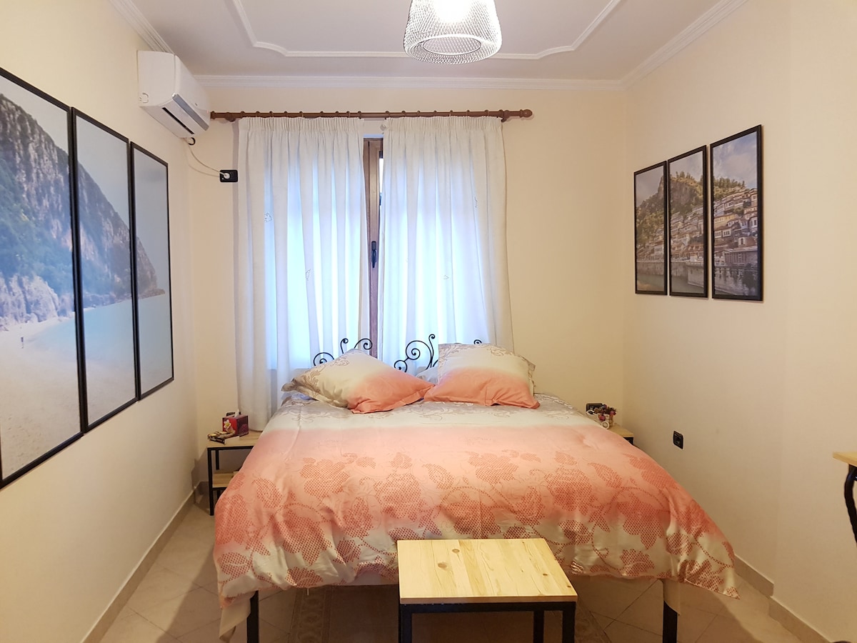 The bedroom features a spacious bed with light pink and floral bedding, flanked by two small side tables. Large windows with sheer curtains allow daylight to enter. The walls are adorned with framed photographs, while an air-conditioning unit is mounted above the bed.
