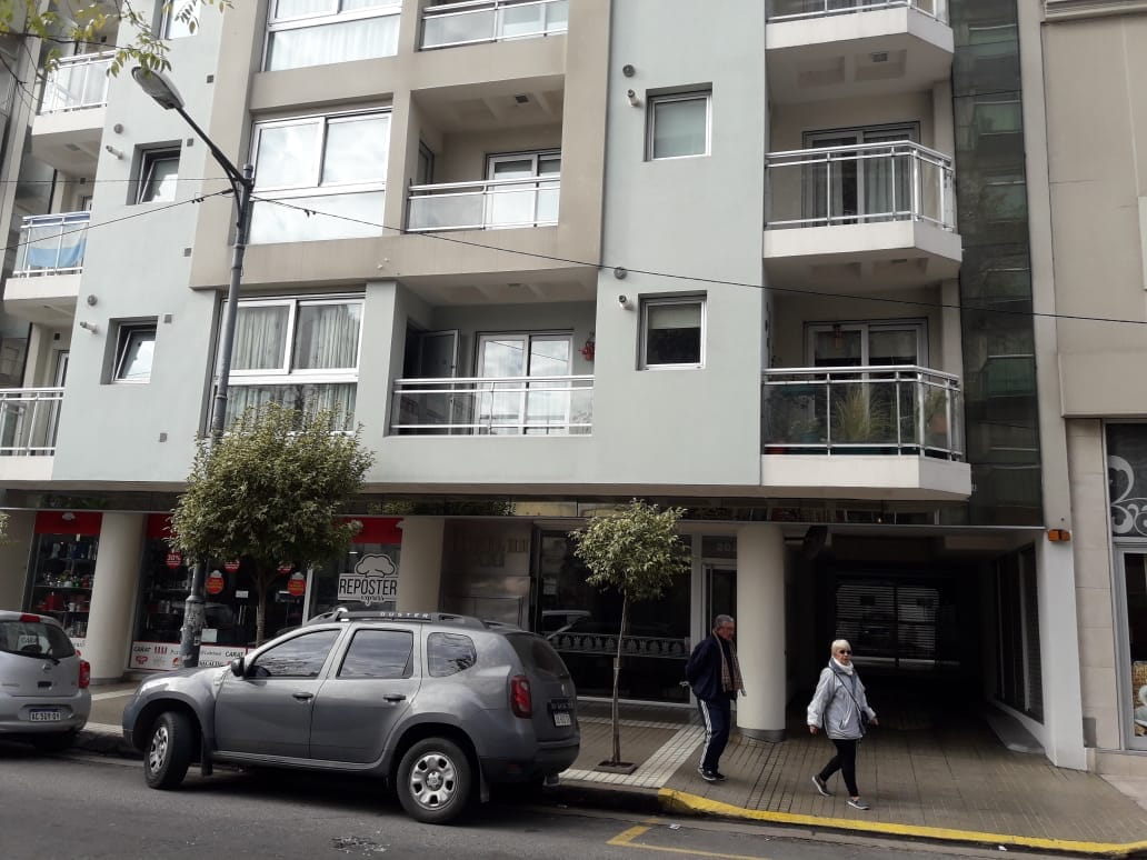 The modern building features a clean architectural design with multiple balconies on the upper floors. A welcoming entrance is visible, along with parked vehicles on the street. Trees are positioned along the walkway, contributing to the urban landscape.