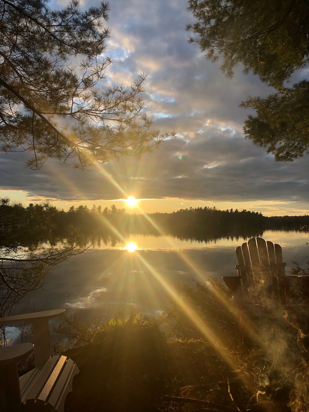 A serene lakeside view features the sun setting over calm waters, casting reflections on the surface. In the foreground, a wooden chair is positioned for lounging, framed by trees and soft clouds. The scene captures a moment of tranquility and natural beauty.