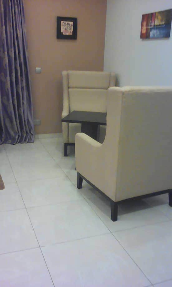 A small seating area features two light-colored armchairs positioned around a dark wooden table. A wall art piece hangs above, with a soft purple curtain draped to the left, contrasting with the light tiled floor and beige walls.