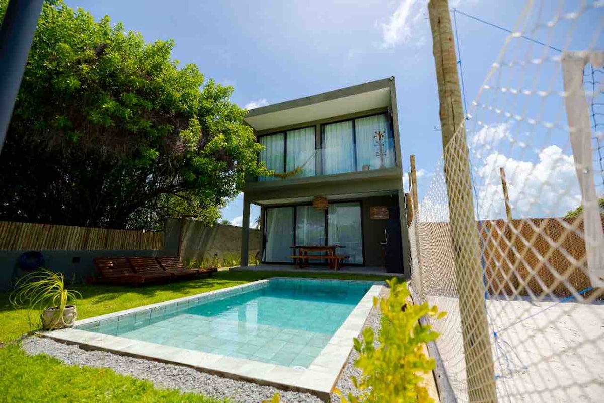 A modern two-story house is presented, featuring large glass doors that open to an inviting pool area. The surrounding yard is well-maintained, with a grassy lawn and a shaded seating area. A white netted fence encloses a separate play area, enhancing the outdoor space.