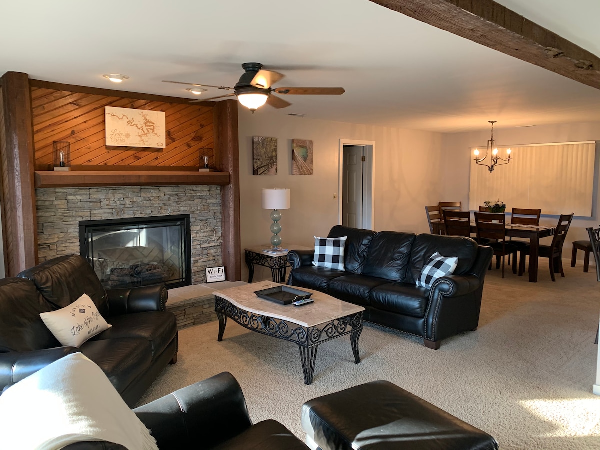 A spacious living area features a stone fireplace with a wooden mantle, flanked by comfortable black leather sofas. A coffee table rests in the center, while a dining table with dark wooden chairs is visible in the background. Warm lighting creates an inviting atmosphere.