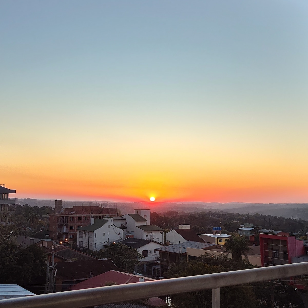 A vibrant sunset is captured over the horizon, displaying hues of orange and yellow blending into a blue sky. Below, rooftops of nearby buildings create a varied landscape, with trees lightly framing the view. The setting suggests a serene and picturesque evening.
