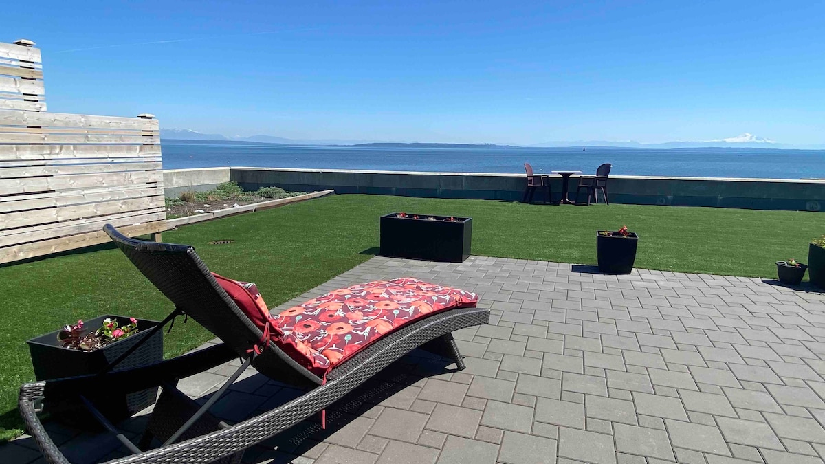 A spacious outdoor patio features a comfortable lounge chair with a red cushion, positioned on green artificial grass. Flower planters are placed around the space, and seating for two is visible, all overlooking a stunning ocean view under a clear blue sky.