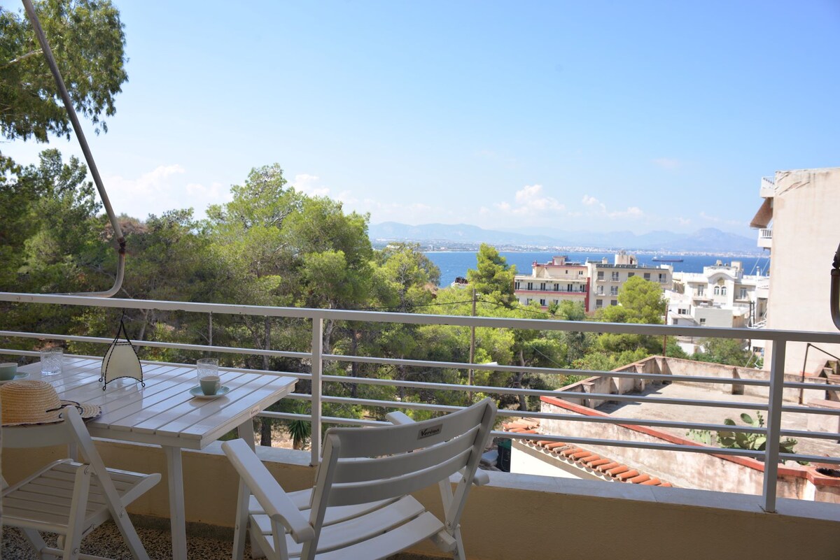 A spacious balcony is shown with a view of the sea and distant mountains. A white table and two chairs are positioned on the balcony, surrounded by greenery. The sky is clear, allowing natural light to enhance the inviting outdoor space.