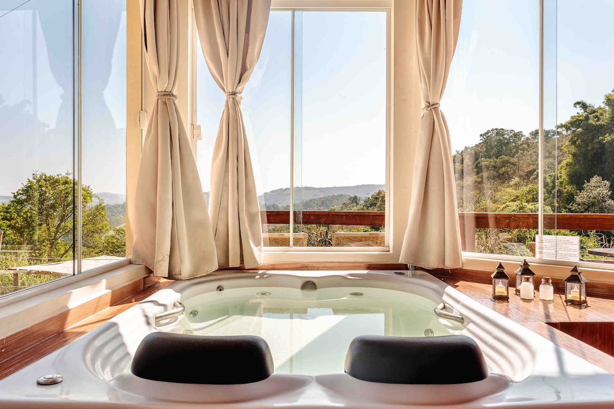 A private jacuzzi with hydro and heating is placed in front of large windows, offering a view of the surrounding nature. Soft curtains frame the scene, while a few decorative lanterns sit nearby, creating a serene atmosphere for relaxation.