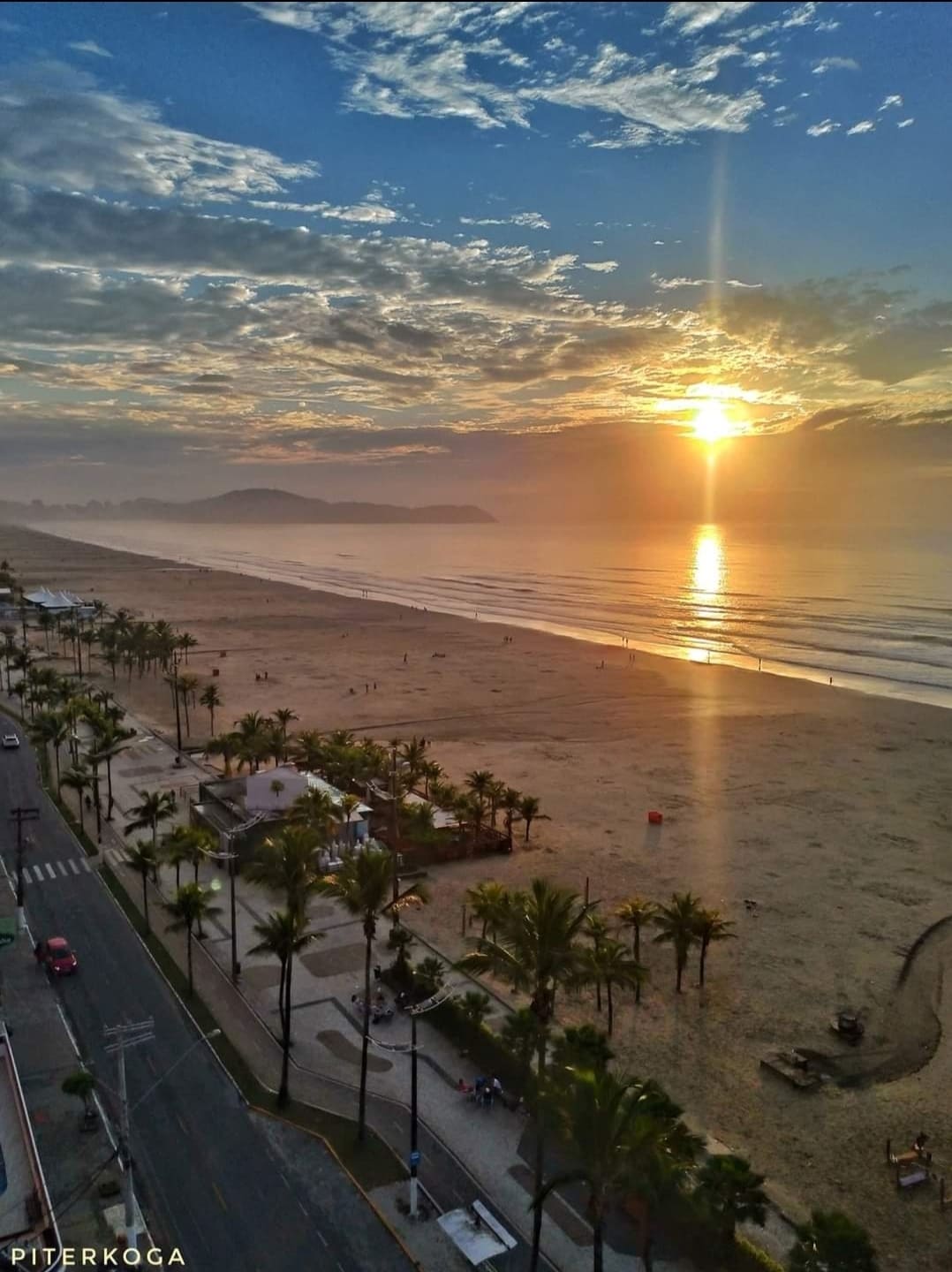 A beach view at sunrise is captured, displaying gentle waves lapping the shore. Silhouetted figures can be observed walking along the sand, while palm trees line the pathway. Soft colors in the sky create a serene backdrop for this coastal landscape.