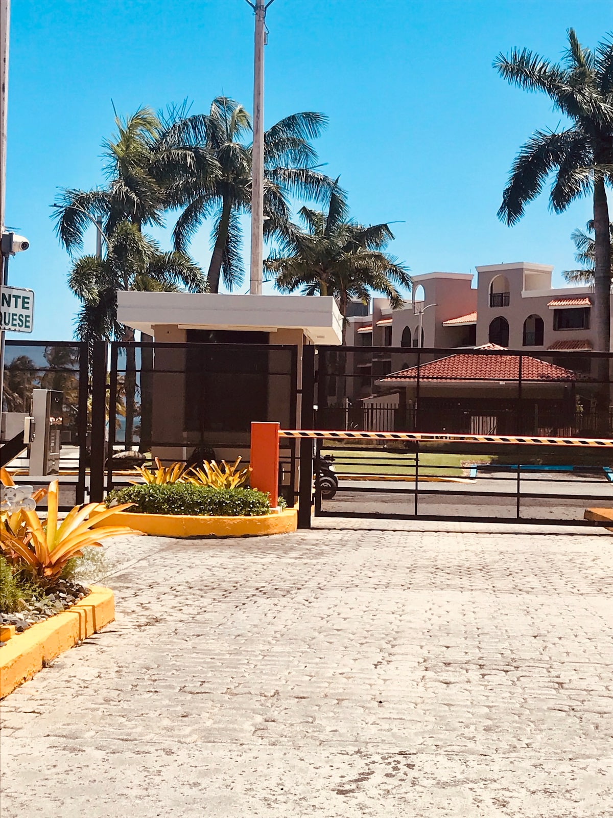 A secure entry gate is visible, framed by vibrant tropical plants and palms. The surrounding area features a well-maintained pathway leading to the condo complex, which is partially seen in the background, showcasing a mix of modern architecture and lush greenery.