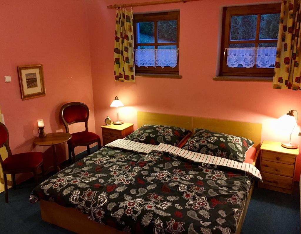 A cozy bedroom features a spacious bed with a patterned duvet cover and two bedside tables, each with a lamp. Two windows dressed with colorful curtains allow natural light, while a small table and chair are positioned against the wall.