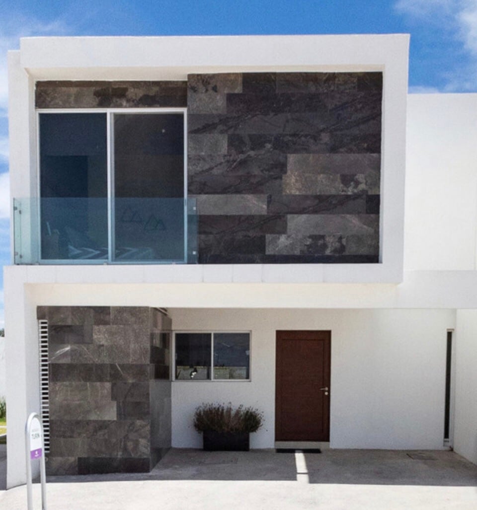 The exterior of the house features a modern design with a combination of dark stone cladding and a glass balcony. A wooden door is visible, along with a small planter next to the entrance. The clear blue sky serves as a backdrop.