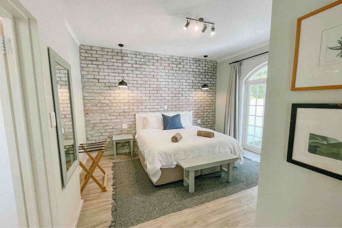 A spacious bedroom features a light-colored bed adorned with a blue accent pillow and neatly arranged towels. Exposed brick walls add texture, while a small table and an elegant mirror complement the layout. Natural light enters through French doors, enhancing the serene ambiance.