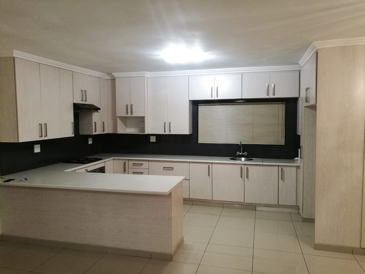 A modern kitchen features light-colored wood cabinetry with a sleek design. Black countertops contrast with the cabinets, while a window allows natural light to enter. An island with a flat surface is positioned centrally, equipped with a stovetop nearby and a sink located under the window.