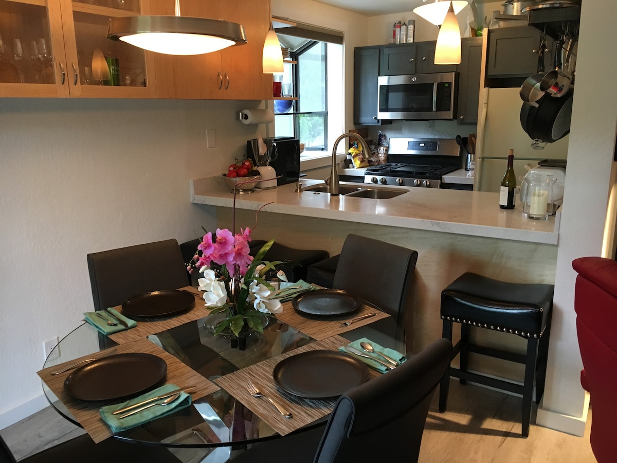 A dining area features a round glass table set for four, surrounded by black chairs. A vase with pink and white flowers is centered on the table. The kitchen is visible in the background, showcasing modern appliances and cabinetry.